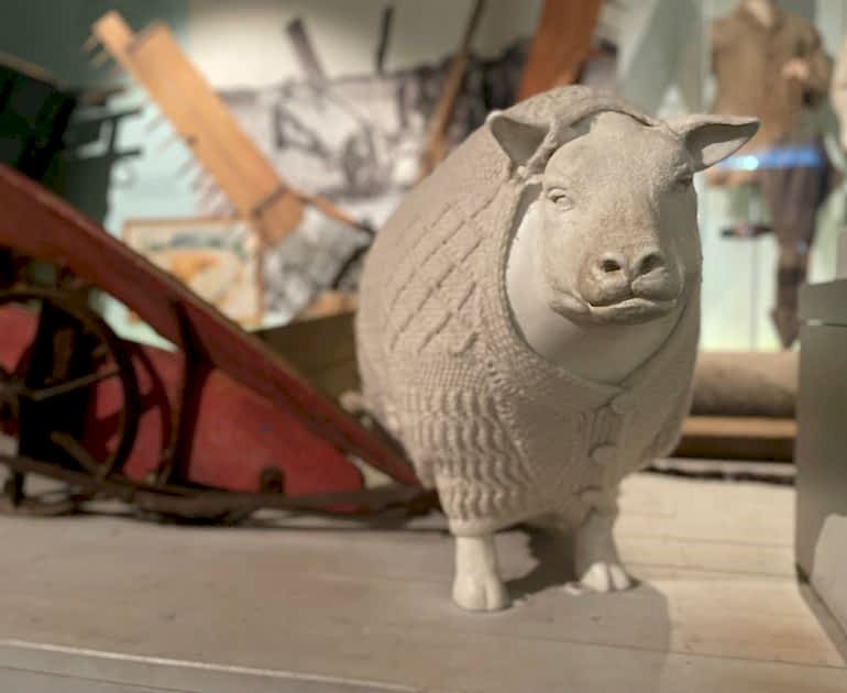 A white plastic sheep wearing a cream-coloured cardigan with dorset buttons, against a backdrop of old agricultural technologies